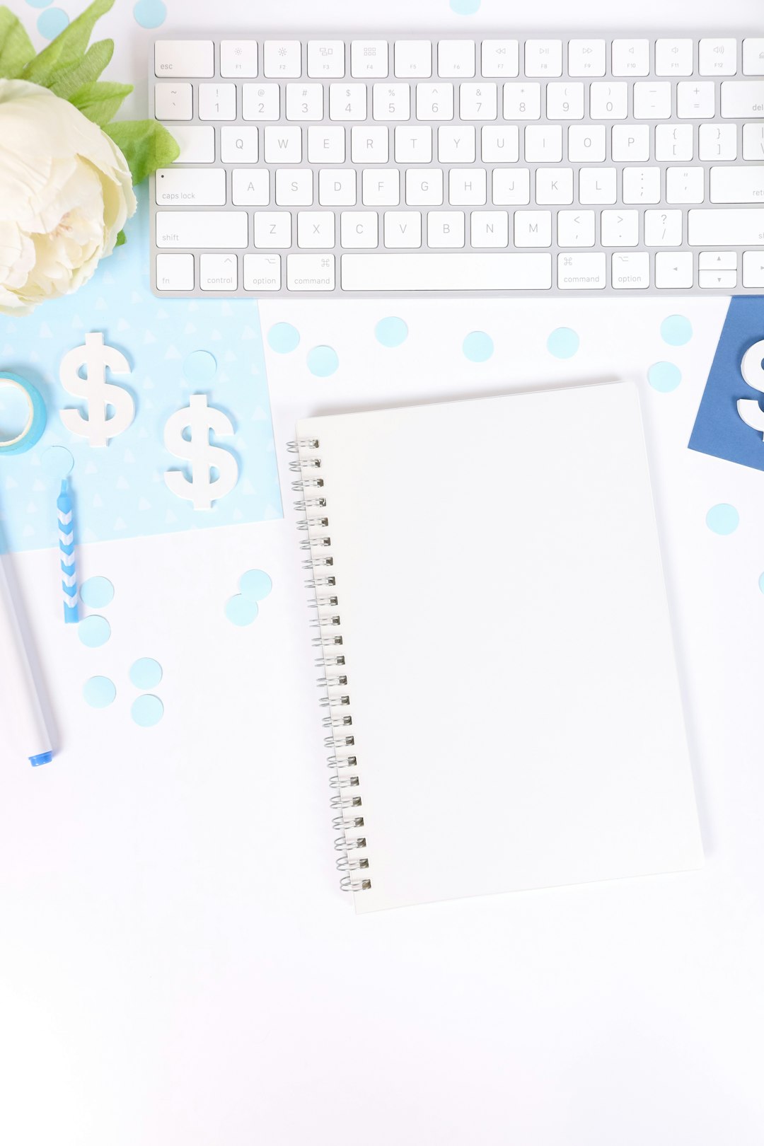 This feminine flat lay styled stock photo features a white desk with light blue and dark blue props like a keyboard, white flowers, markers, confetti, a notebook, and more.